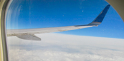 A photo taken through the window of an airliner showing the aircraft's right wing, part of the frame of the window, the azure-blue sky and a solid layer of clouds far below